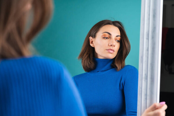 Young woman dressed in blue turtleneck, golf, posing sensually in front of the mirror