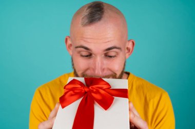 Turkuaz arka planda turuncu tişörtlü sakallı bir adam, neşeli bir hediye kutusuyla, neşe saçar, hoş bir şekilde gülümser.