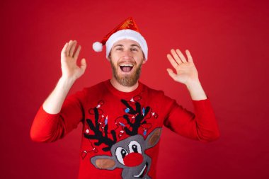 Kırmızı arka planda Noel Baba şapkası ve Noel süveteri giyen kızıl sakallı adam neşeli bir şekilde poz veriyor, heyecanla gülümsüyor.