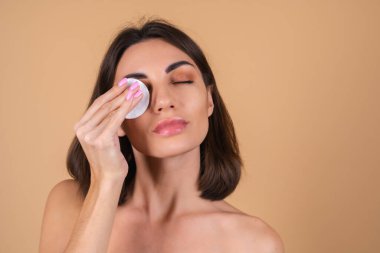 Bej arka planda çıplak omuzları olan, yüzünü temizlemek için sünger tutan, güzellik portresi yapan, cildine özen gösteren üstsüz bir kadının yakın portresi.