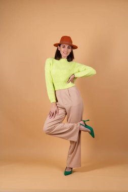 Bej arka planda, pantolonlu, kazaklı, şapkalı ve ayakkabılı tam gelişmiş şık bir kadın.