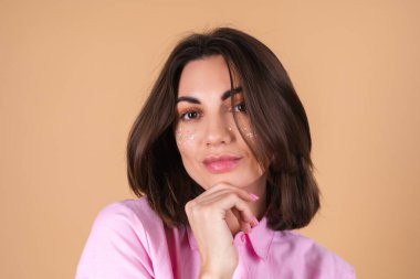 Bej arka planda pembe elbiseli genç bir kadının portresi. Parlak makyajlı ve sevimli küpeleri var. Olumlu, nazik romantik bir poz veriyor.