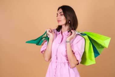 Bej arka planda pembe elbiseli genç bir kadının portresi. Parlak makyajlı. Ayakkabıları ve alışveriş paketleri var.