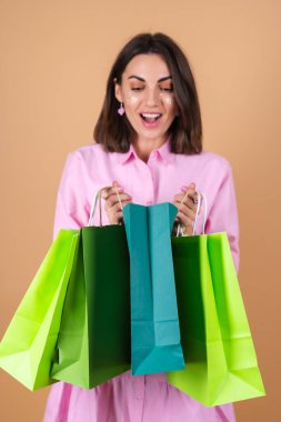 Bej arka planda pembe elbiseli genç bir kadının portresi. Parlak makyaj malzemesi elinde. Güzel alışveriş paketleri.
