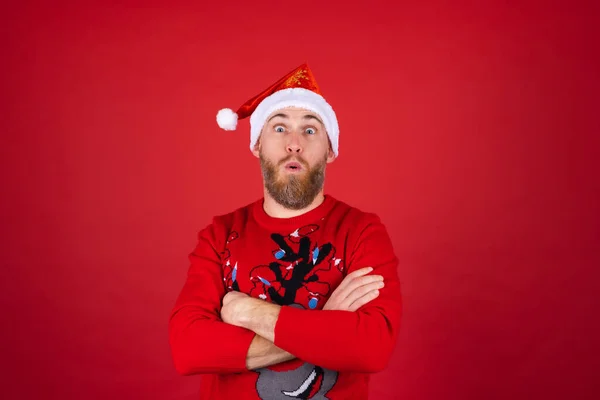 赤い背景にあるサンタの帽子とクリスマスセーターの赤い髭の男は陽気にポーズをとって 興奮して笑顔 — ストック写真