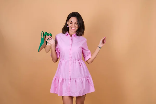 若いです女性上のベージュの背景にピンクのドレスでキラキラメイクアップは緑の靴を保持しています 低ヒールポンプポジティブにポーズ かわいいです浮気笑顔 — ストック写真