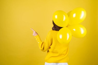 Sarı kazaklı bir kızın sırtından bir çerçeve, elinde balonlar, neşeli bir ruh hali, bir doğum günü partisi beklentisi içinde, parmağını sola gösteriyor.