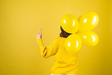 Sarı kazaklı bir kızın sırtından bir çerçeve, elinde balonlar, neşeli bir ruh hali, bir doğum günü partisi beklentisi, bir parmağını havaya kaldırır.