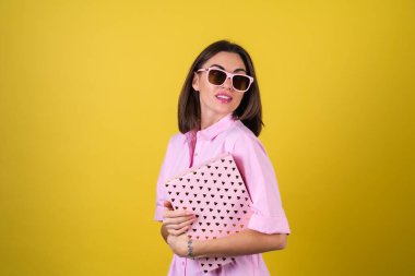 Pembe elbiseli ve sarı arka planda gözlüklü şık bir genç kadın elinde hediye kutlamasıyla en iyi bayramı, doğum gününü, sevgililer gününü kutluyor.