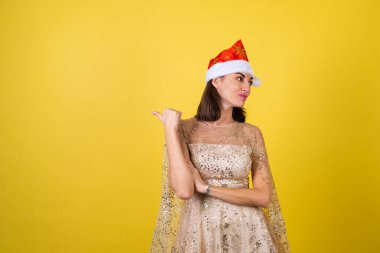 Yeni yıl arifesinde genç bir kadın parlak parlak bej bir elbise ve Noel Baba şapkası bayramları kutluyor, parlak pembe ruj dudak parlatıcısı bir parmağını sola gösteriyor.