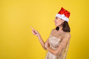 Yeni yıl arifesinde genç bir kadın parlak parlak bej bir elbise ve Noel Baba şapkası bayramları kutluyor, parlak pembe ruj dudak parlatıcısı bir parmağını sola gösteriyor.