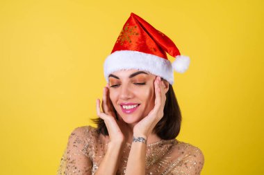 Yeni yıl arifesinde genç bir kadın parlak parlak bej bir elbise ve Noel Baba şapkası bayramları kutluyor, parlak pembe ruj dudak parlatıcısı bir parmağını sola gösteriyor.