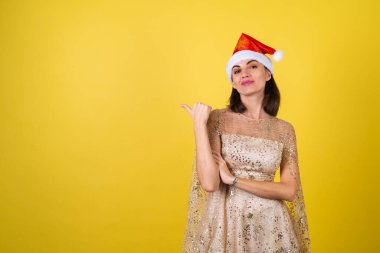Yeni yıl arifesinde genç bir kadın parlak parlak bej bir elbise ve Noel Baba şapkası bayramları kutluyor, parlak pembe ruj dudak parlatıcısı bir parmağını sola gösteriyor.