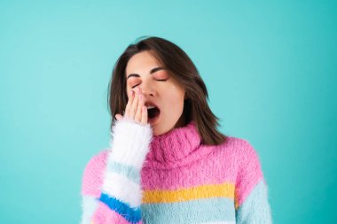 Mavi arka planda parlak renkli süveter giyen genç bir kadın uykulu gözler, tatlı esnemeler, uykusuzluk.