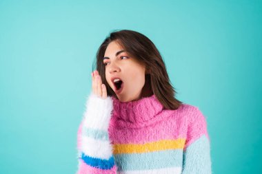 Mavi arka planda parlak renkli süveter giyen genç bir kadın uykulu gözler, tatlı esnemeler, uykusuzluk.