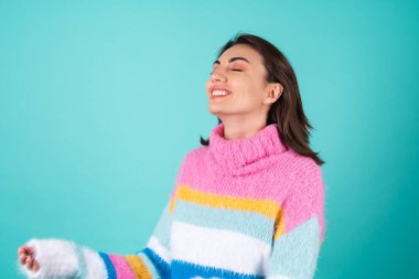 Mavi arka planda parlak renkli süveter giyen genç bir kadın gülümsüyor, kapalı gözlerle anın tadını çıkarıyor.