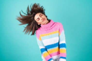 Mavi arka planda parlak renkli bir süveter giyen genç bir kadın. Gözleri kapalı gülümseyen, pürüzsüz saçlı, güzel uçan, dolgun dudaklı, çıplak makyajlı.