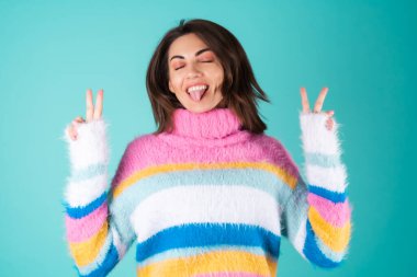 Mavi arka planda parlak renkli süveter giyen genç bir kadın. Sevimli gülümseyen, şakacı, dil gösteren ve jest yapan.