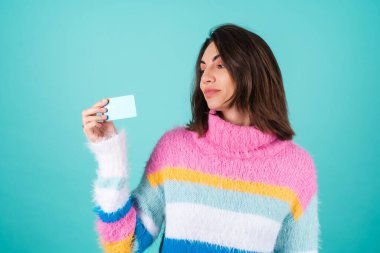 Mavi arka planda parlak renkli süveter giyen genç bir kadın gülümseyen bir kredi kartı gösteriyor.