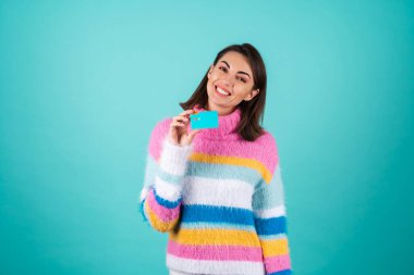 Mavi arka planda parlak renkli süveter giyen genç bir kadın gülümseyen bir kredi kartı gösteriyor.