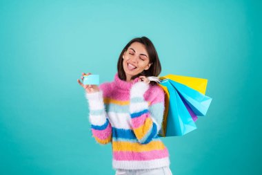 Mavi arka planda parlak renkli bir süveter giyen genç bir kadın bir kredi kartı ve alışveriş torbası tutuyor, mutlu bir şekilde gülümsüyor.
