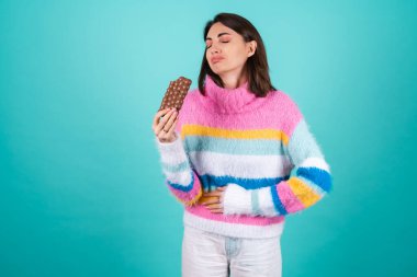 Mavi arka planda parlak renkli süveter giyen genç bir kadın hazımsızlık ve karın ağrısından muzdarip, elinde sütlü çikolatayla