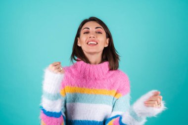 Mavi arka planda parlak renkli bir süveter giyen genç bir kadın, gülümsüyor, neşeyle hareket ediyor, tek başına dans ediyor.