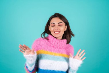 Mavi arka planda parlak renkli bir süveter giyen genç bir kadın, gülümsüyor, neşeyle hareket ediyor, tek başına dans ediyor.