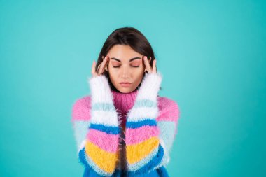 Mavi arka planda parlak renkli süveter giyen genç bir kadın baş ağrısı çekiyor, parmaklarını şakaklarına bastırıyor.