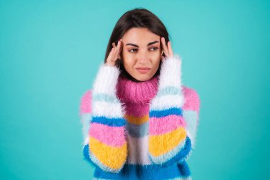 Mavi arka planda parlak renkli süveter giyen genç bir kadın baş ağrısı çekiyor, parmaklarını şakaklarına bastırıyor.
