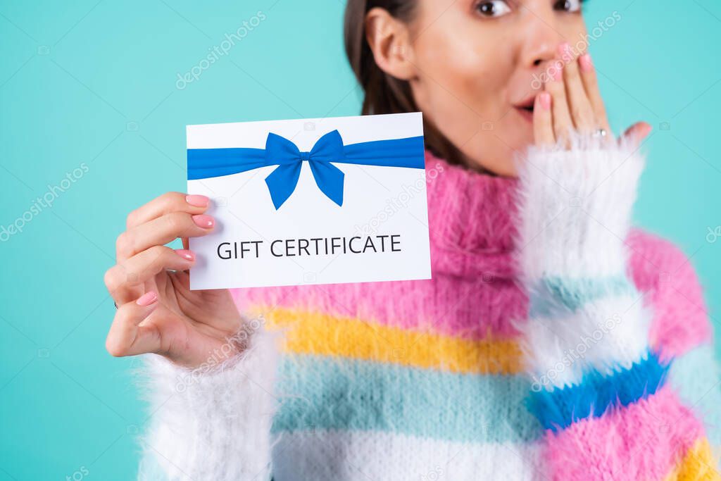 Young woman in a bright multicolored sweater on a blue background holds a gift certificate, smiles enthusiastically, beautiful makeup, nude plump lips