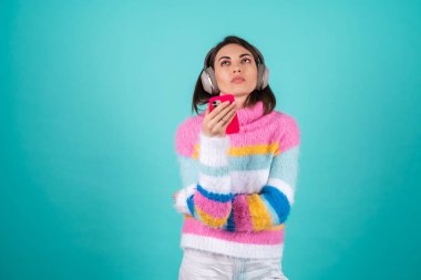 Mavi arka planda parlak renkli bir süveter giyen, kulaklıkları çıkartan genç bir kadın sıkılmış bir ifadeyle telefonu tutuyor.