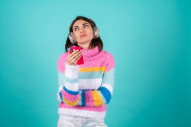 Mavi arka planda parlak renkli bir süveter giyen, kulaklıkları çıkartan genç bir kadın sıkılmış bir ifadeyle telefonu tutuyor.