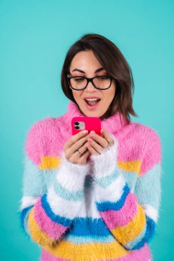 Mavi arka planda, parlak renkli bir süveter giyen genç bir kadın. Gözlüklü, elinde telefon tutan, ağzı açık bir şekilde ekrana bakan.