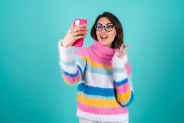 Mavi arka planda, parlak renkli bir süveter giyen genç bir kadın. Gözlüklü, elinde telefon tutan, selfie çeken, kendi fotoğrafını çeken, eğlenen.