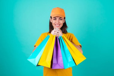 Genç kadın teslimatçı, kurye, mavi arka planda turuncu sarı şapkalı, çok renkli alışveriş poşetleri taşıyor, sipariş veriyor.