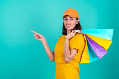 Genç kadın teslimatçı, kurye, mavi arka planda turuncu sarı şapkalı, çok renkli alışveriş poşetleri taşıyor, sipariş veriyor.