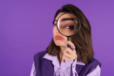 Young woman in shirt and vest on purple background having fun with magnifying glass in hand