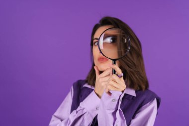 Young woman in shirt and vest on purple background having fun with magnifying glass in hand