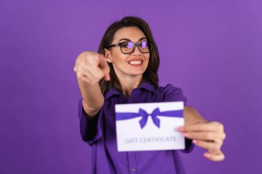 Young woman in a shirt on a purple background with a gift certificate smiling cheerfully, with makeup, lipstick on her lips and glasses, excited clipart