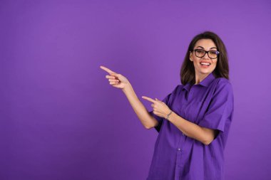 Young woman in a shirt with short sleeves on a purple background, with makeup and stylish glasses, points a finger to the left with a smile