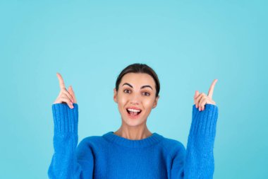 Beauty portrait of a young woman in a blue knitted sweater and natural day makeup, smiling cheerfully and pointing her finger up to an empty space