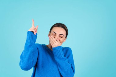 Beauty portrait of a young woman in a blue knitted sweater and natural day makeup, smiling cheerfully and pointing her finger up to an empty space