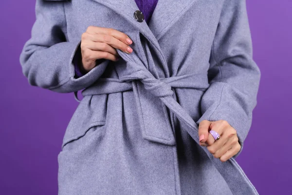 Woman's hands with a big ring on the finger tying the belt of a gray winter coat on a purple background