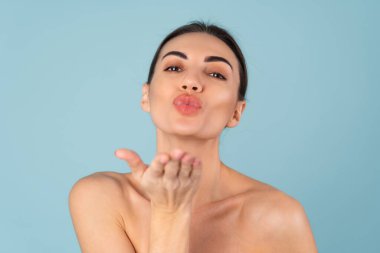 Close beauty portrait of topless woman with perfect skin and natural make-up, plump nude lips, on a blue background, sends a kiss