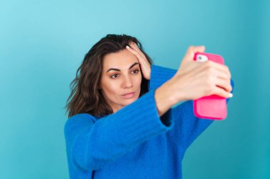 Mavi örgü süveterli ve doğal makyajlı bir kadın, güçlü bir yüz, cep telefonunda parlak pembe kapaklı kendi fotoğrafını çekiyor.