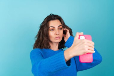 Mavi örgü süveterli ve doğal makyajlı bir kadın, güçlü bir yüz, cep telefonunda parlak pembe kapaklı kendi fotoğrafını çekiyor.