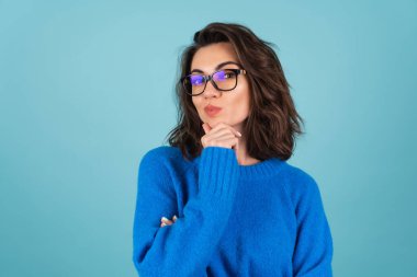 Mavi örgü kazaklı, doğal makyajlı, kısa kıvırcık saçlı, gözlerinde gözlük olan bir kadın, düşünceli bir şekilde kameraya bakıyor, meditasyon yapıyor.