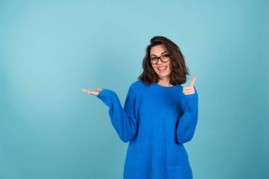 Mavi örgü kazaklı bir kadın, kısa kıvırcık saçlı, gözlüklü, neşeli bir şekilde gülümseyen, boş bir yeri işaret eden, avucunda tutan