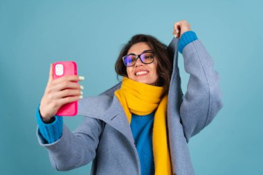 Mavi örgü süveter, gözlük, sarı parlak atkı ve gri ceketli bir kadının bahar sonbahar portresi, bir telefon tutuyor ve kendi fotoğrafını çekiyor.